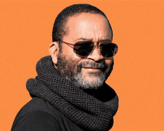 A person wearing aviator vintage sunglasses and a black knit scarf against an orange background.