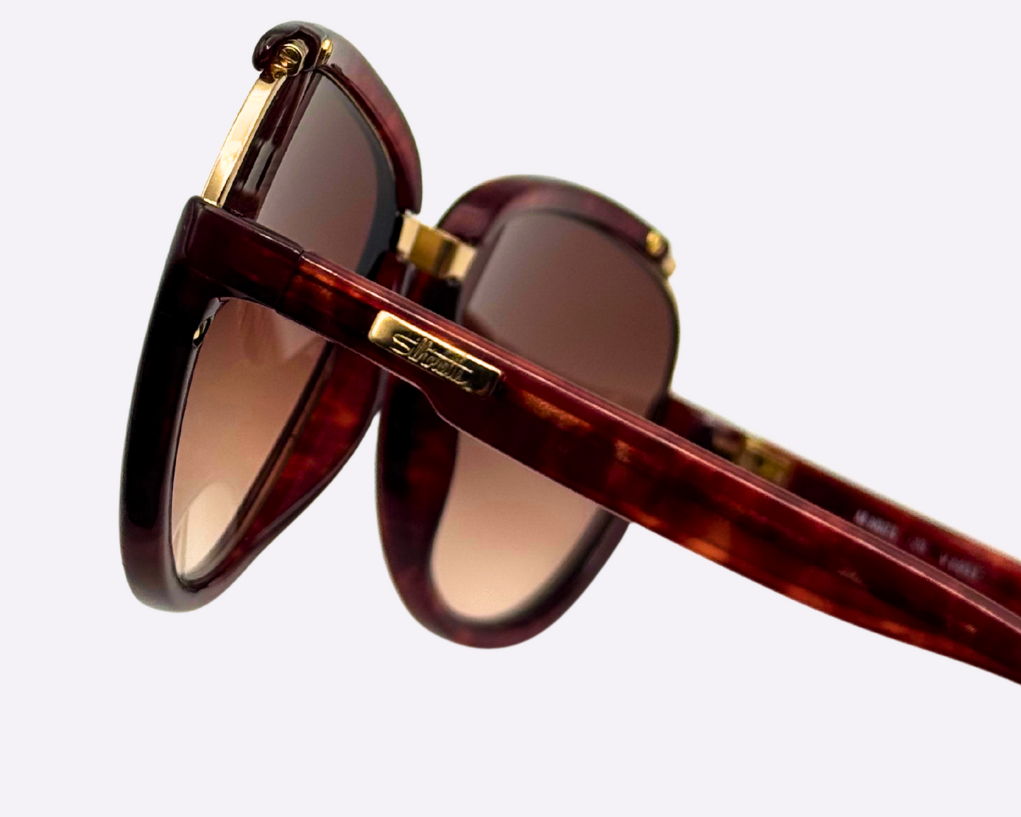 Close-up of the temple and Silhouette logo on vintage 1980s M8023/20 cat-eye sunglasses with a gold and tortoise optyl and metal frame and original brown gradient lenses, shown on a light gray background.