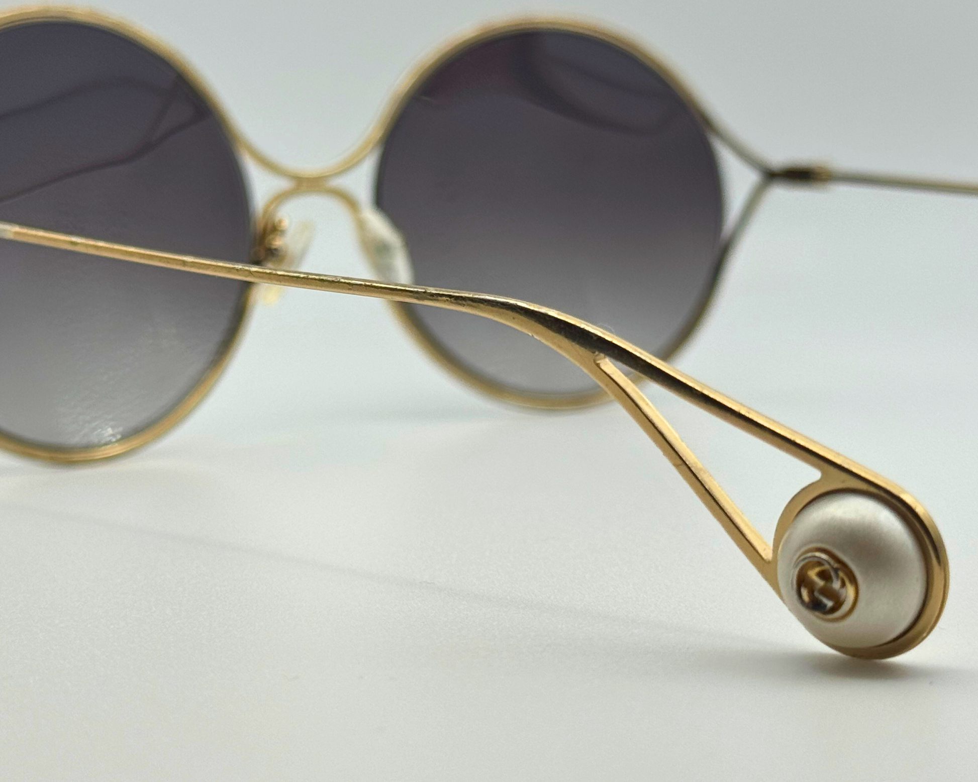 Close-up of the temple and pearl-like logo on gold-framed Gucci sunglasses, shown on a light gray background.